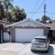 A silver car is parked in front of a small house with a white garage door and driveway, surrounded by lush greenery.