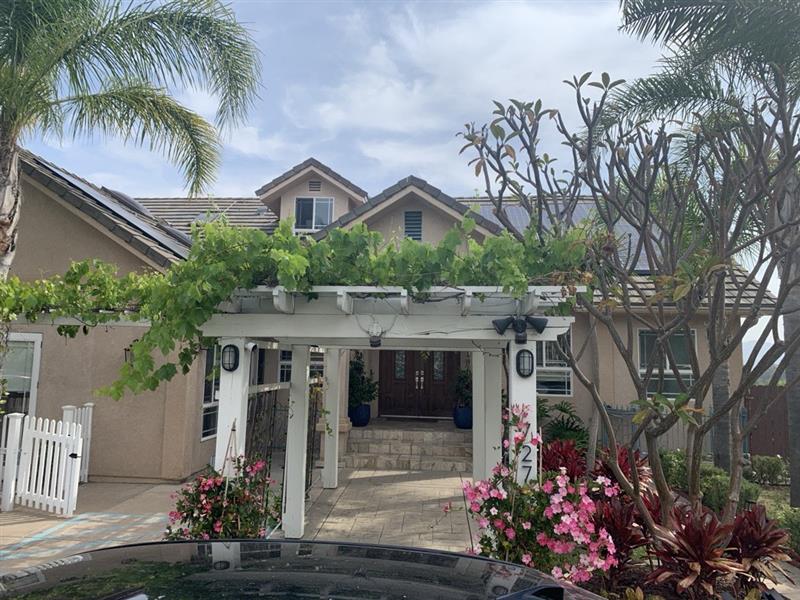 A charming house with a vine-covered pergola, lush tropical plants, and a white picket fence under a partly cloudy sky.