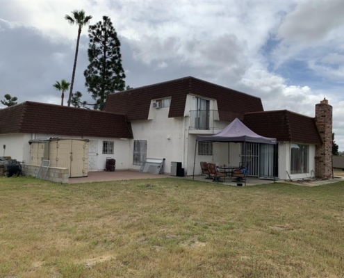 2ec747c2-02e2-4ba2-b5f8-c5cf6cea9251 A white house with a brown roof, patio furniture, and a cozy shed sits on a grassy lawn under cloudy skies, shaded by a stylish canopy.
