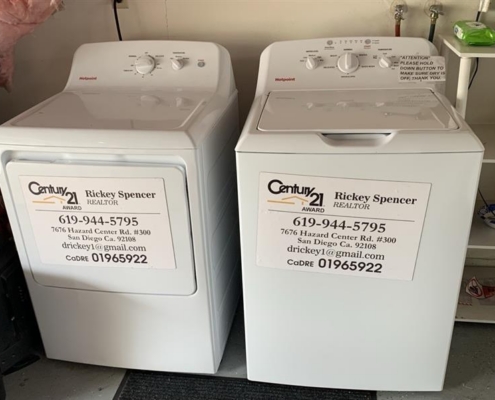 db30a08a-90ec-49f3-9515-2574e3fe6129 Two white laundry machines with Century 21 realtor signs taped to the front are set up in a bright laundry room.