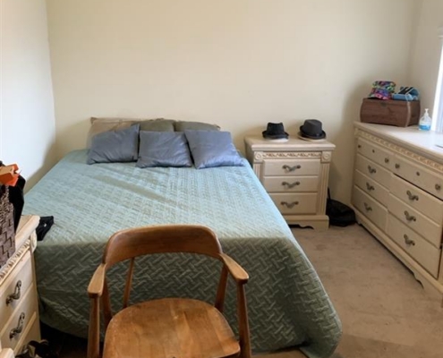 e664656f-9eae-427f-a2aa-15f556303bf5 Small bedroom with a neatly made bed, three pillows, dressers, and a stylish wooden chair at the foot of the bed.