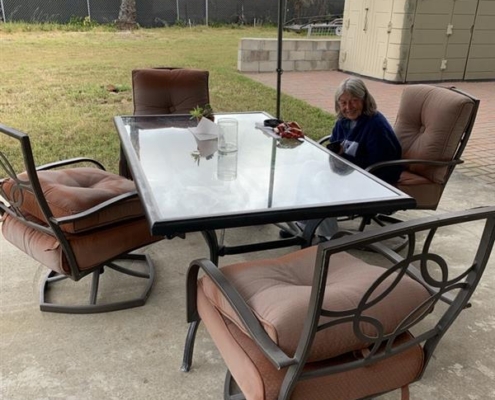 f2c49010-81b1-4338-8f64-0e8daaf4c3cc A woman sits by a glass patio table with four cushioned chairs on a concrete slab in a spacious backyard.