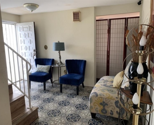 f7ccde9b-c2e5-4eb4-bba9-9f3348ba9b85 Bright entryway with patterned tile floor, blue chairs, a lamp, and a cozy small sofa near an open door and modern stairs.